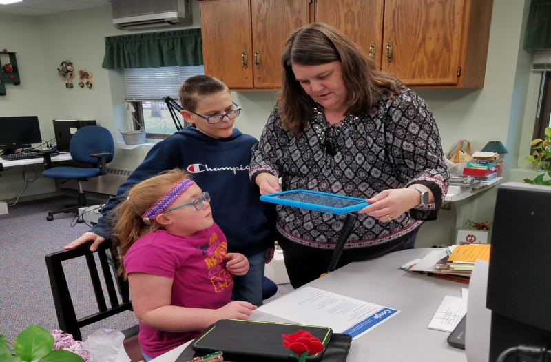 Laurie stands holding an iPad while 2 students (one seated and one standing) watch and wait, looking at the iPad.