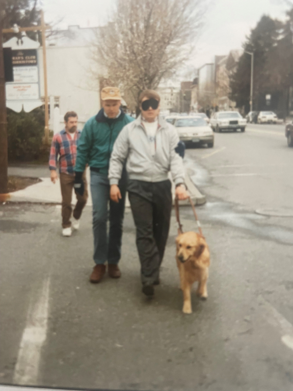 Paul wearing a blindfold and holding onto the leash of a guide dog while training to become an O&M specialist. There are two people behind him observing him as he walks down a sidewalk.