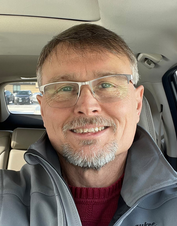 A close-up image of Paul, taken as a selfie in a car. He has graying short hair and facial hair and is wearing glasses and a gray zippered sweatshirt.
