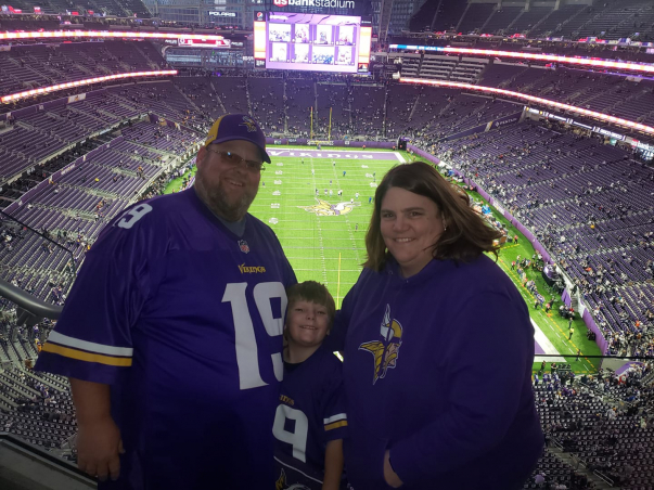 Laurie, her husband Lee and son Eli wear Vikings gear and stand smiling in the stands of the Vikings stadium. The football field can be seen behind them.