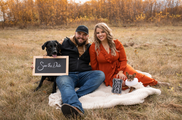 Lilly is seated next to her fiance Zach on the ground with their dogs. A sign states Save the Date, 6/3/2023, for their wedding date.  