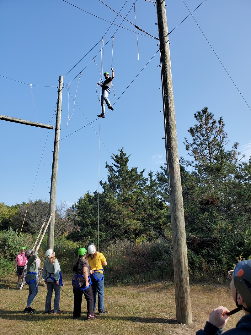 Elias is standing on a wire high above the ground and holding onto another wire above his head. A few people can be seen on the ground below. He is wearing a helmet and a harness.