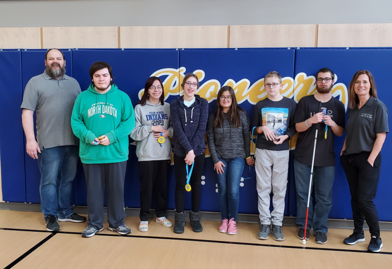 Ryan stands in a line with 6 students and Mrs. Williams in the gym in South Dakota during a goalball tournament there.